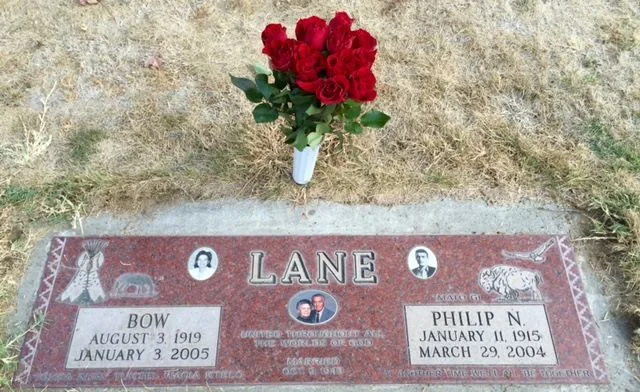 Red granite grave marker for Bow Lane (August 3, 1919 – January 3, 2005) and Philip N. Lane (January 11, 1915 – March 29, 2004), with photos, tipi and buffalo carvings, and a vase of red roses.