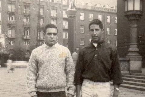 Sepia photograph of two young men standing in a Munich city square in 1962, Phil Lane Jr. and his high school friend Mike Friedman, with tall buildings and an ornate lamppost behind them.