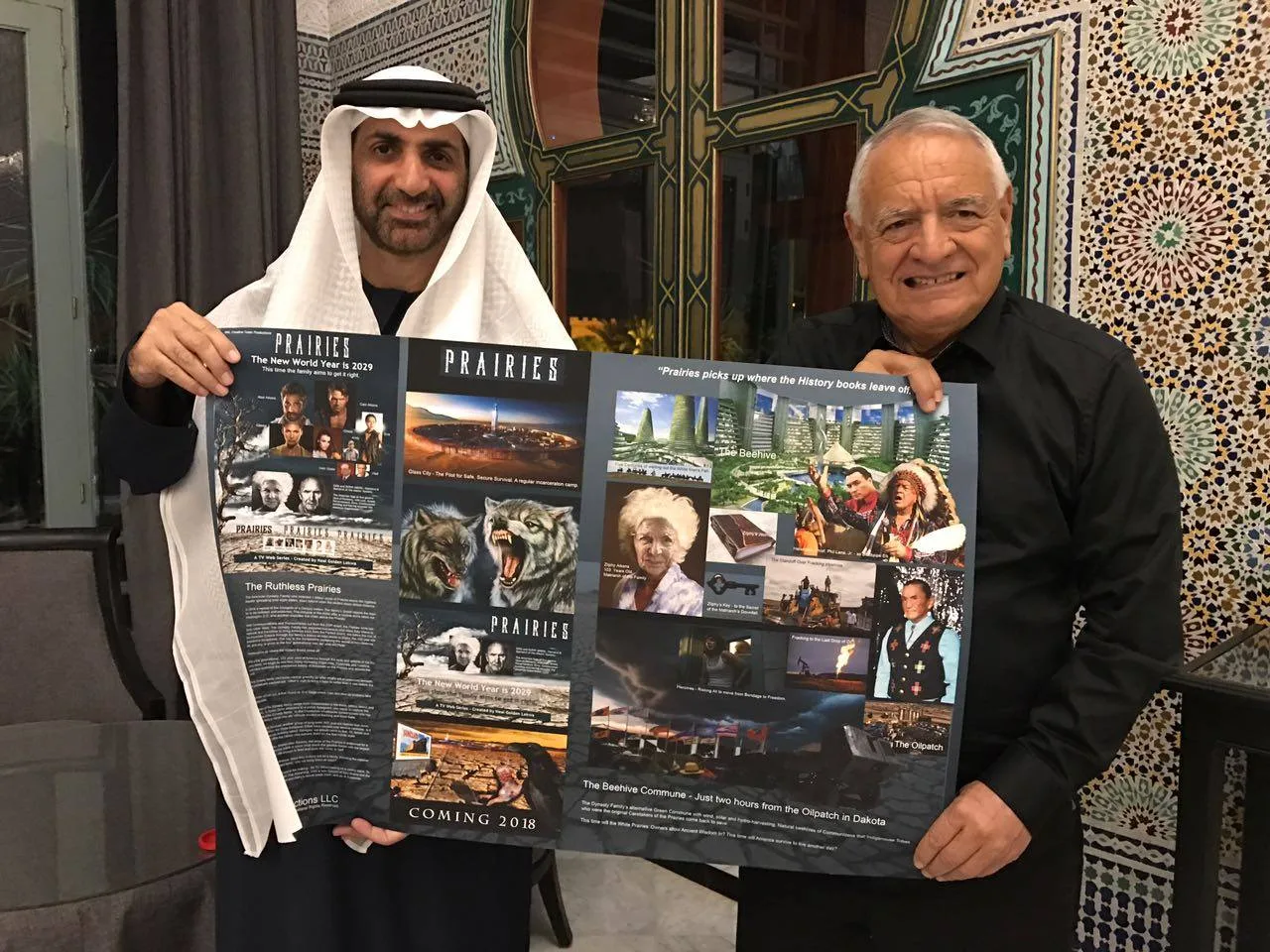 Sheikh Abdul Aziz, in a white headscarf and dark robe, and Phil Lane Jr., in a black shirt, hold up a large poster for the Prairies TV series in a tiled room.
