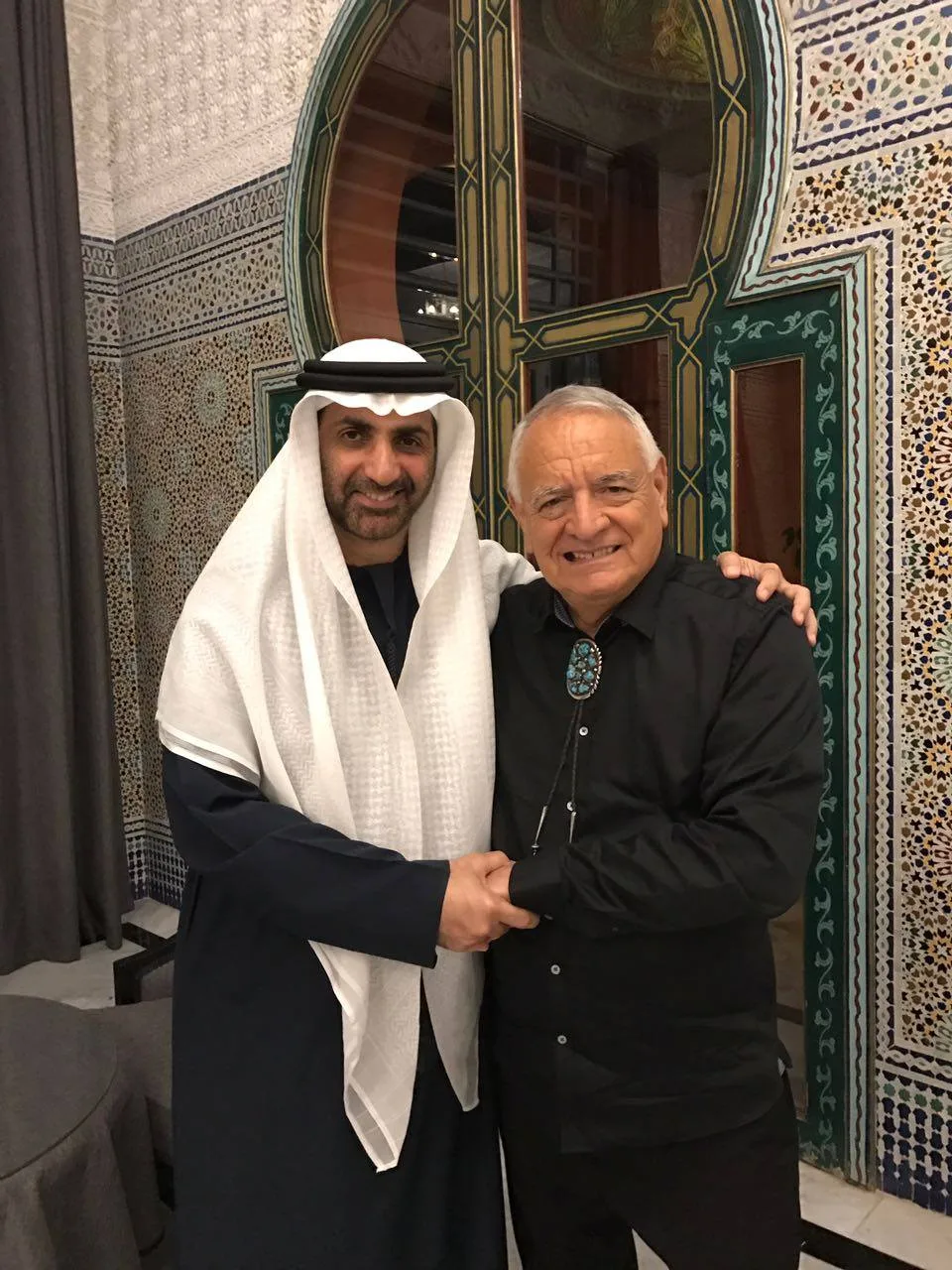 Sheikh Abdul Aziz and Phil Lane Jr. smile, clasping hands with arms around each other, before a green arched doorway and colorful Moroccan mosaic tiles.