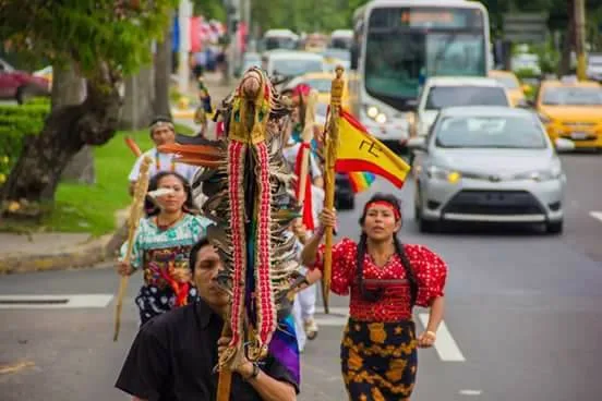 Indigenous runners in traditional dress carry feathered staffs and a flag down a city street, with cars and a bus following behind.