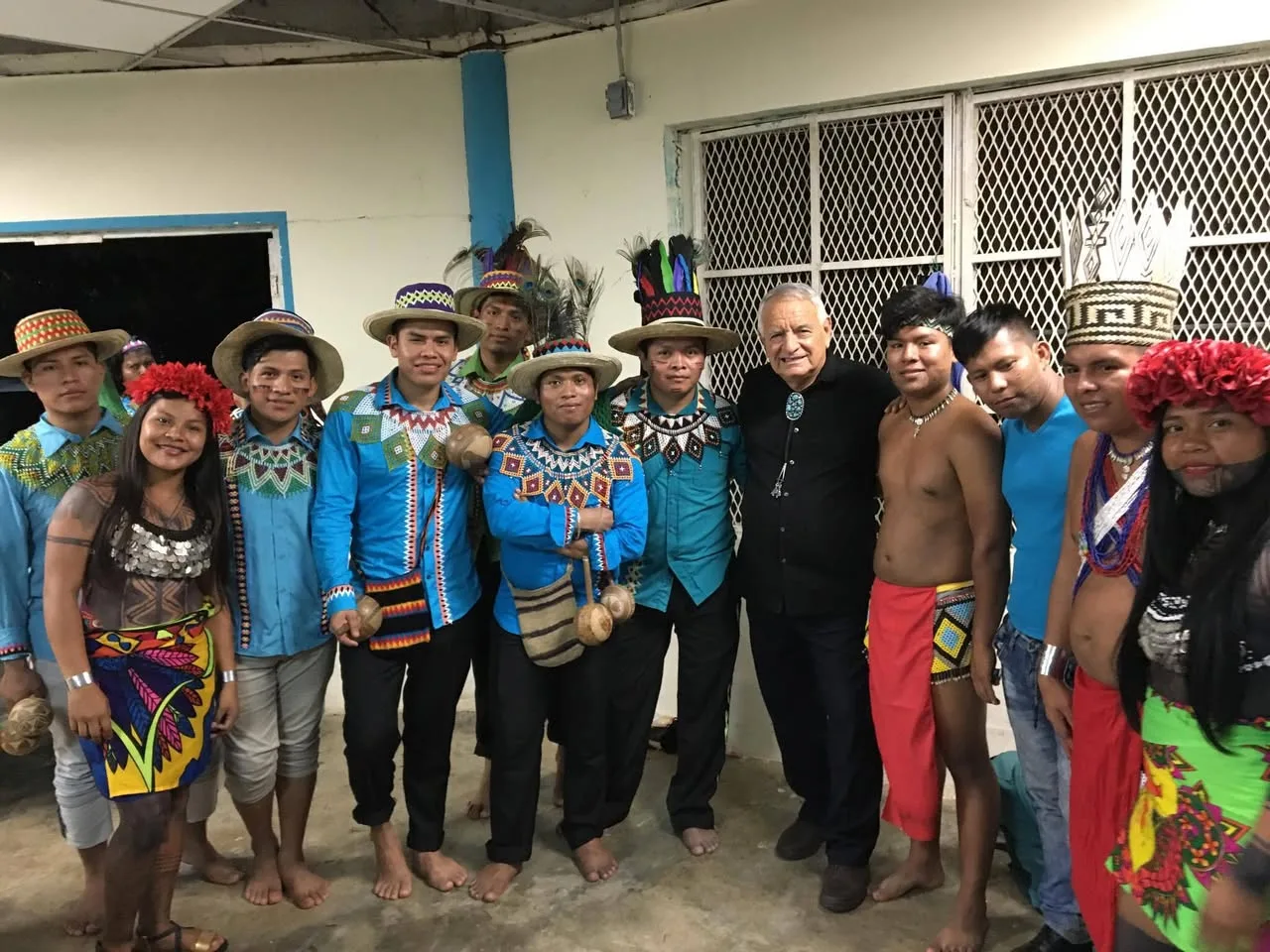 Phil Lane Jr., an older man in a black shirt and bolo tie, stands with young Embera and Ngabe Bugle people in traditional dress: beaded collars, woven hats, feathers and painted skirts.