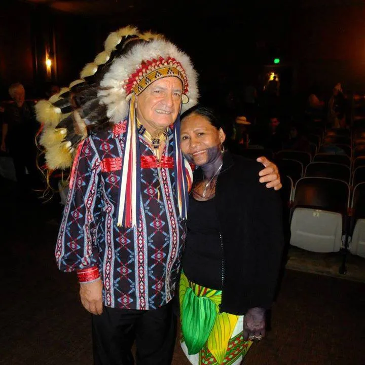 Phil Lane Jr., in a feather headdress and patterned shirt, embraces Sister Irma Caisamo Hermenet, who wears a black top and bright green and yellow skirt, in a theater.