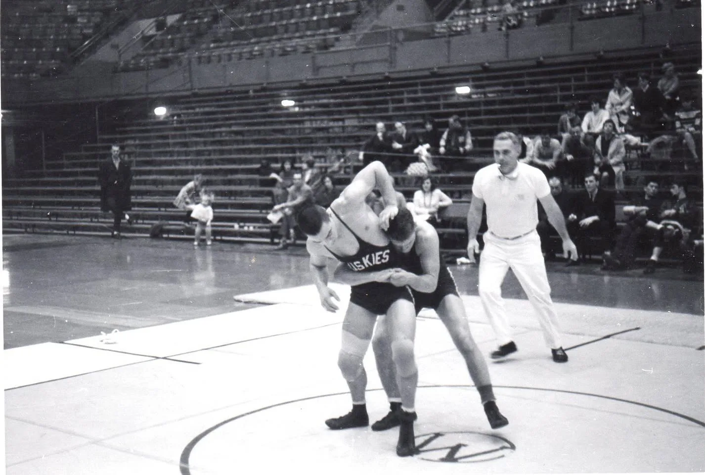 Black-and-white photo of two wrestlers grappling on a mat marked with a W in a university gym, one wearing a Huskies singlet, as a referee in white watches and spectators sit in the stands.