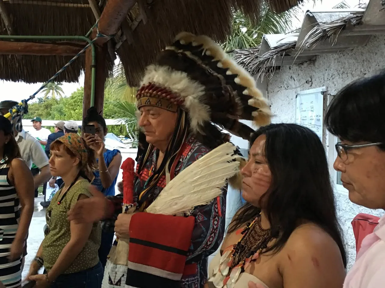 An elder in a large feather headdress, holding an eagle feather fan and a red-wrapped staff, stands beneath a thatched roof among Indigenous women and onlookers.
