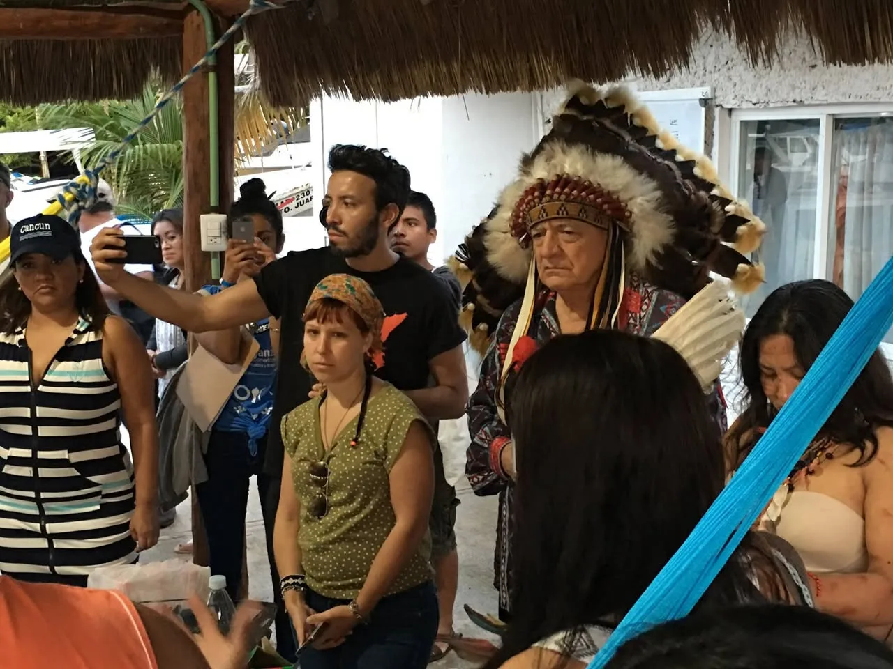 An elder in a feather headdress stands with a gathering under a thatched shelter while people around him record the ceremony on their phones.