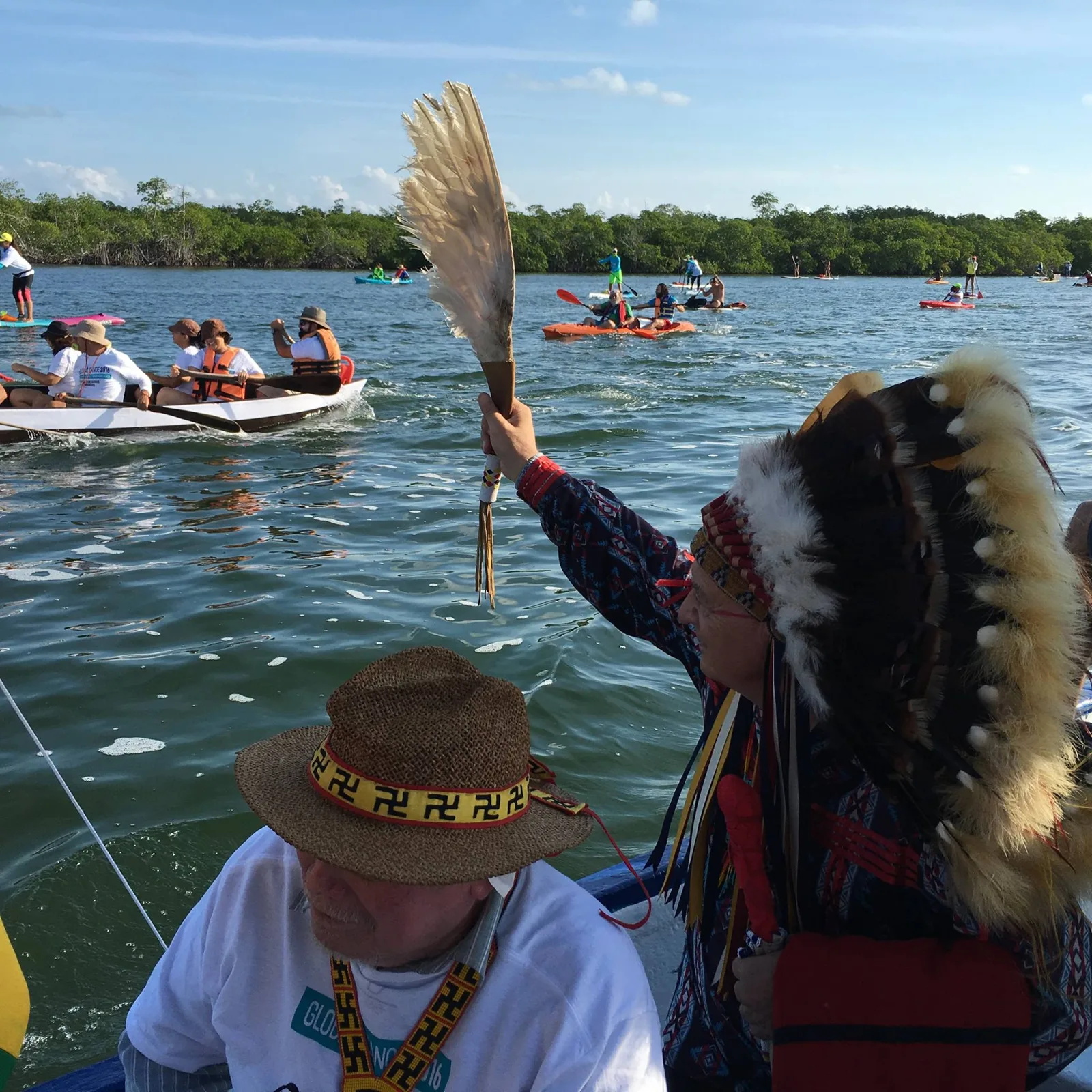 An elder in a feather headdress raises an eagle feather fan from a boat as canoes, kayaks and paddleboards move across water lined with mangroves.