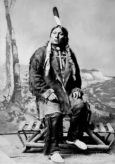 Historic studio portrait of a Hunkpapa chief seated on a rustic bench, wearing a fringed buckskin coat, a large cross pendant and a single feather in his hair.