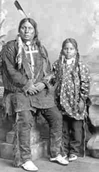 Historic studio portrait of a Lakota chief seated with a cross pendant and feather, beside a young girl with long braids in a patterned dress.