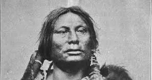 Close-up historic portrait of a Lakota man with long hair, earrings and a fur robe, looking steadily at the camera.
