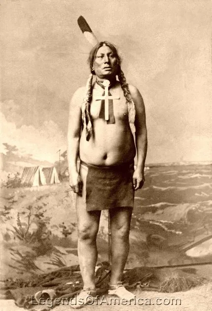 Sepia studio portrait of a standing Lakota chief, bare-chested in a breechcloth, wearing a large cross pendant and a feather, before a painted camp backdrop.