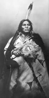 Historic portrait of a Lakota chief with a feather in his hair, wrapped in a decorated robe and holding a round feathered object.