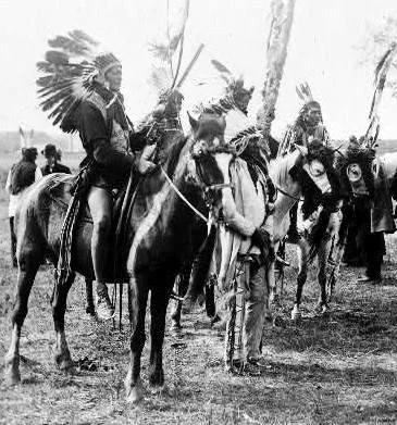Historic photograph of Lakota men in feathered war bonnets on horseback and standing beside decorated horses in an open field.
