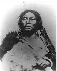 Historic portrait of a Lakota man with long hair and a stern expression, wrapped in a fur robe.