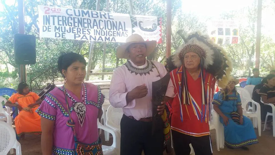 Elsy M. Pedrol Flores in a pink Ngäbe dress, Chief Marcos Samudio in a straw hat holding a carved torch, and an elder in a feather headdress, under a banner for the 2nd Intergenerational Summit of Indigenous Women of Panama.