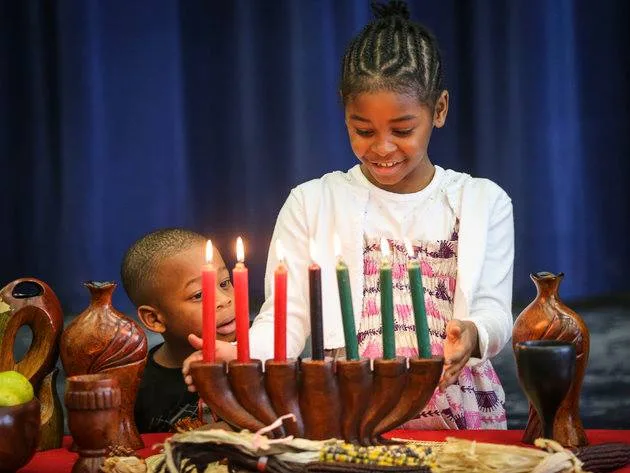 A smiling girl and a young boy beside a wooden kinara holding red, black and green candles, surrounded by carved wooden vases and harvest items on a red cloth.