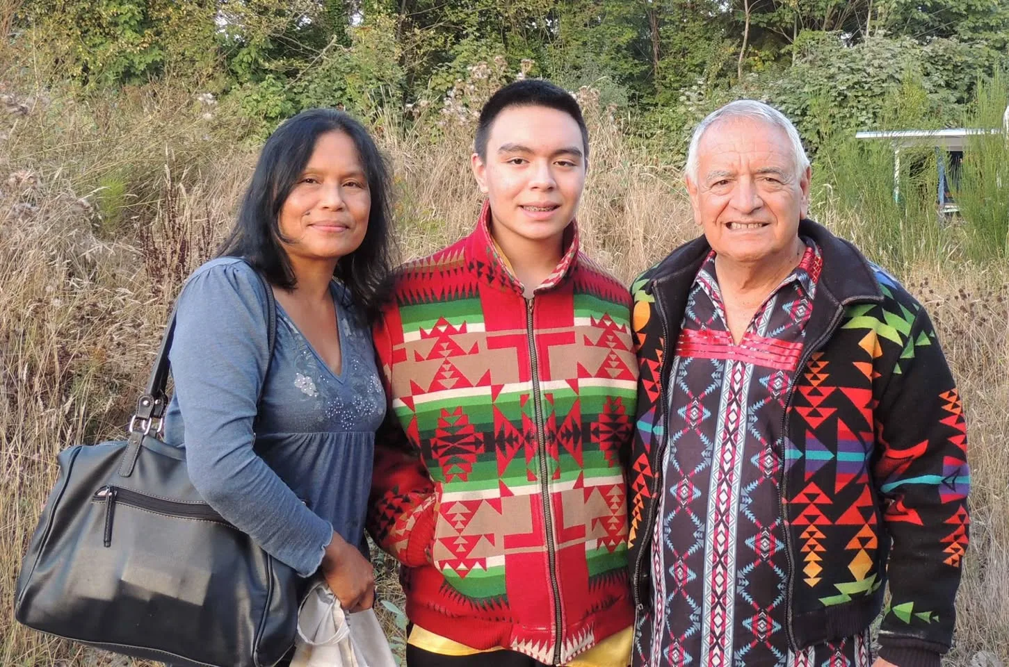 A smiling woman with a shoulder bag, a teenage boy in a red and green patterned jacket, and a white-haired elder in a colorful geometric jacket stand together before tall grass and trees.