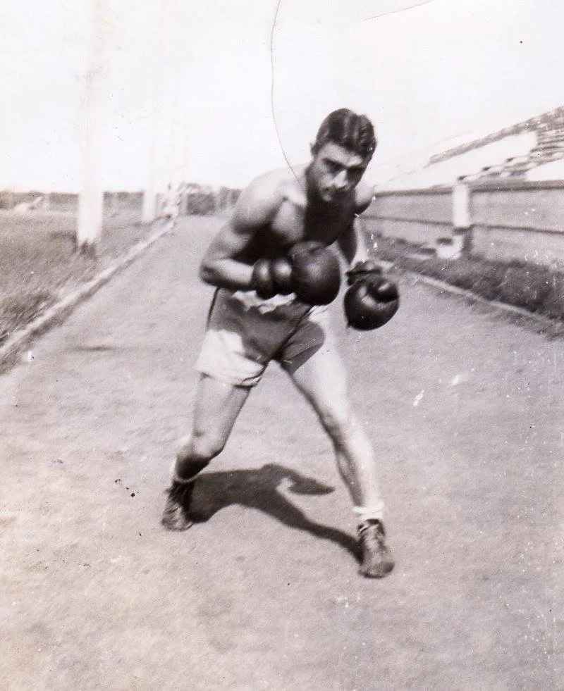 Old photo of a young shirtless boxer in gloves and shorts crouched in a fighting stance on a dirt path.