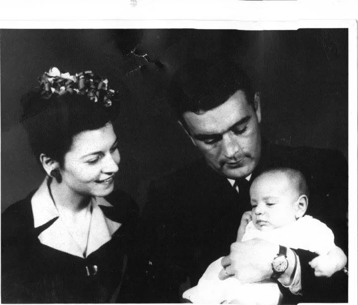 Black and white portrait of a smiling woman and a man in a suit holding a baby in a white gown.