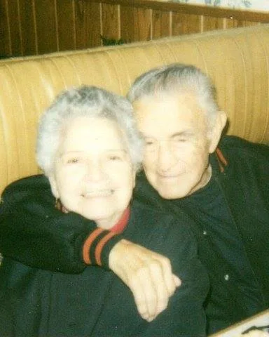 An elderly couple with white hair embrace and smile while seated in a restaurant booth.