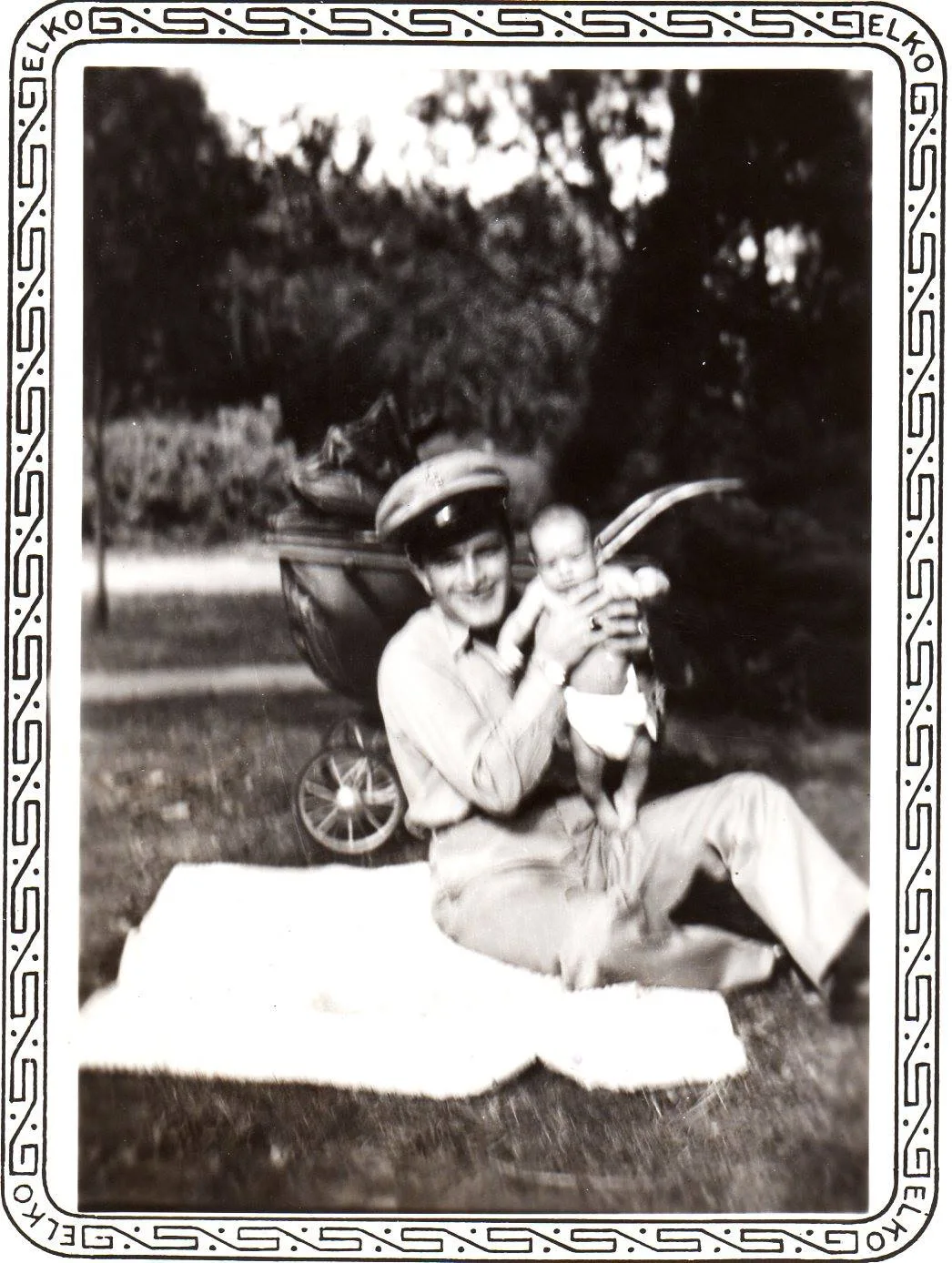 Old photo of a young man in a cap holding up a baby while sitting on a blanket on the grass beside a baby carriage.