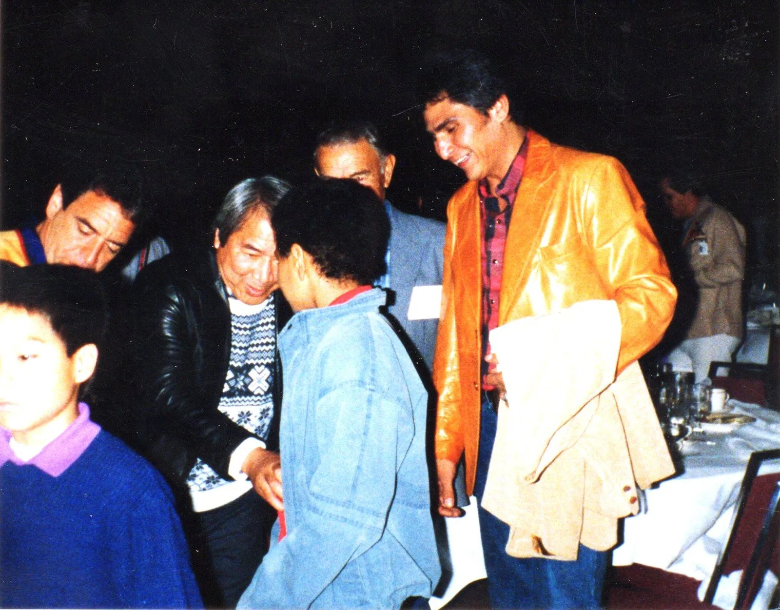 Several men and boys greet one another warmly at a banquet; a man in an orange leather jacket smiles at a boy in denim.