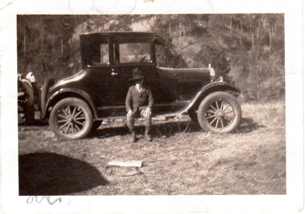 Old photograph of a small boy in a hat and suit seated on the running board of a vintage car.