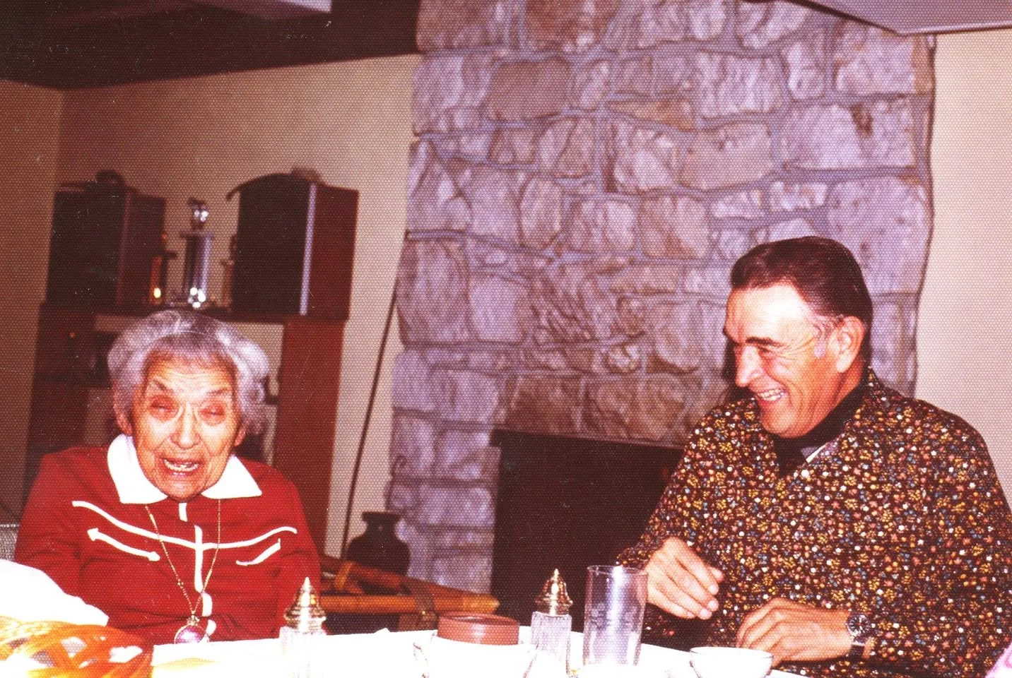 An elderly woman in red laughs beside a smiling man in a patterned shirt at a table in front of a stone fireplace.