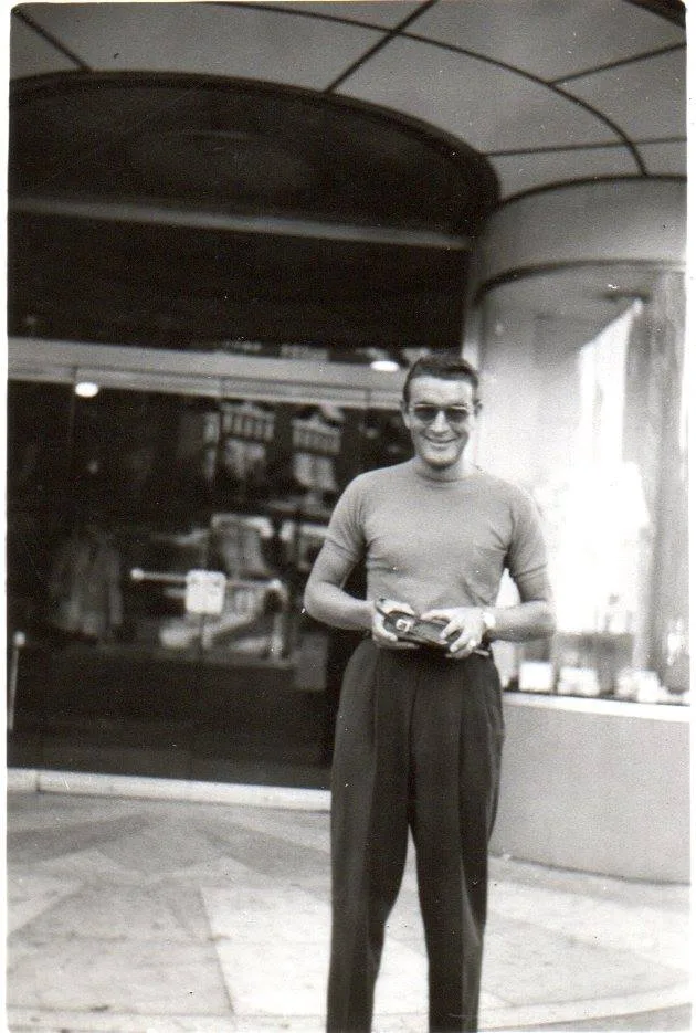 Old photo of a smiling young man in sunglasses and a T-shirt holding a camera outside a storefront.