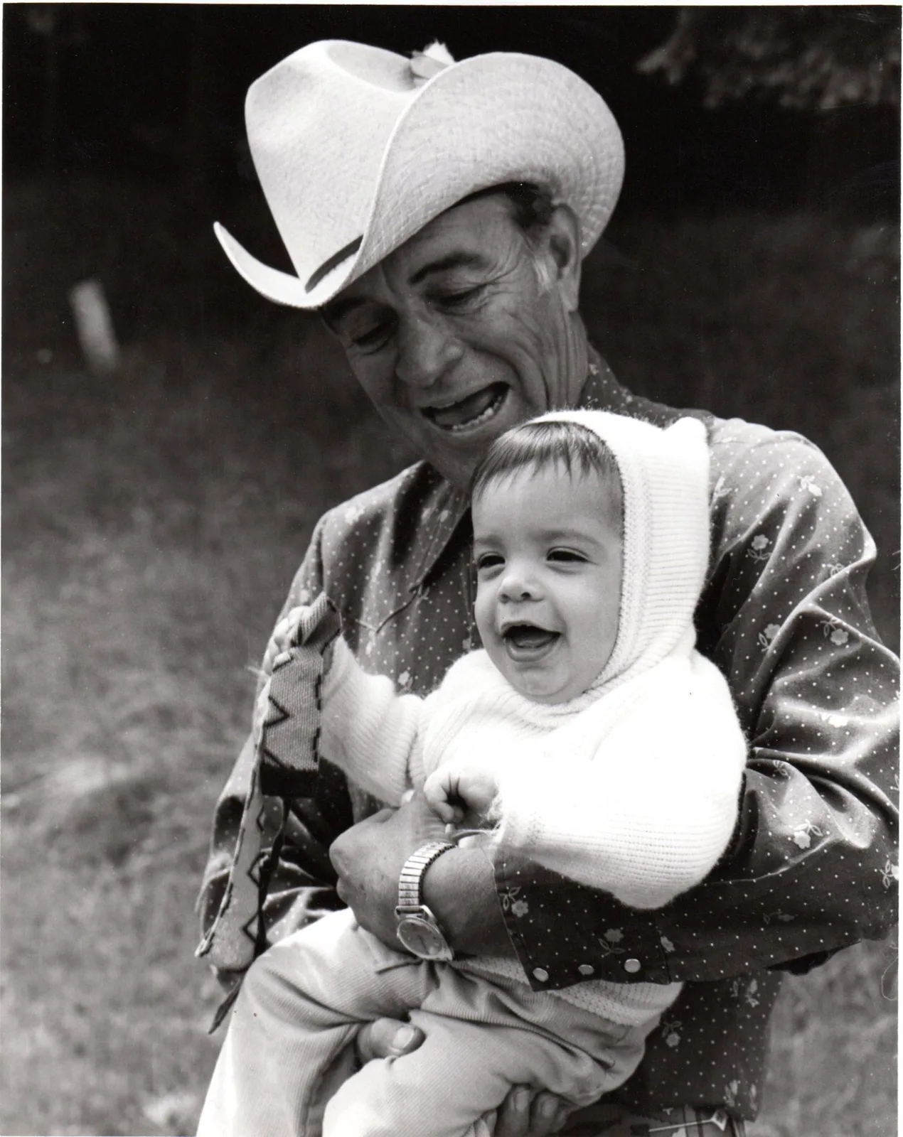 Black and white photo of a man in a cowboy hat laughing as he holds a giggling baby in a white knit hood.