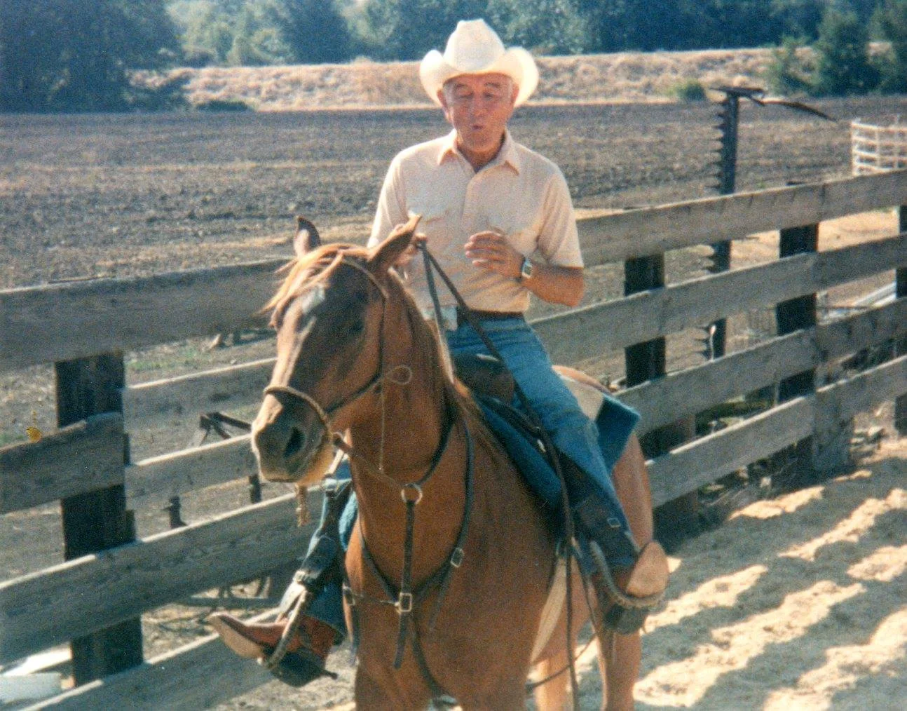 An older man in a white cowboy hat rides a chestnut horse along a wooden corral fence.