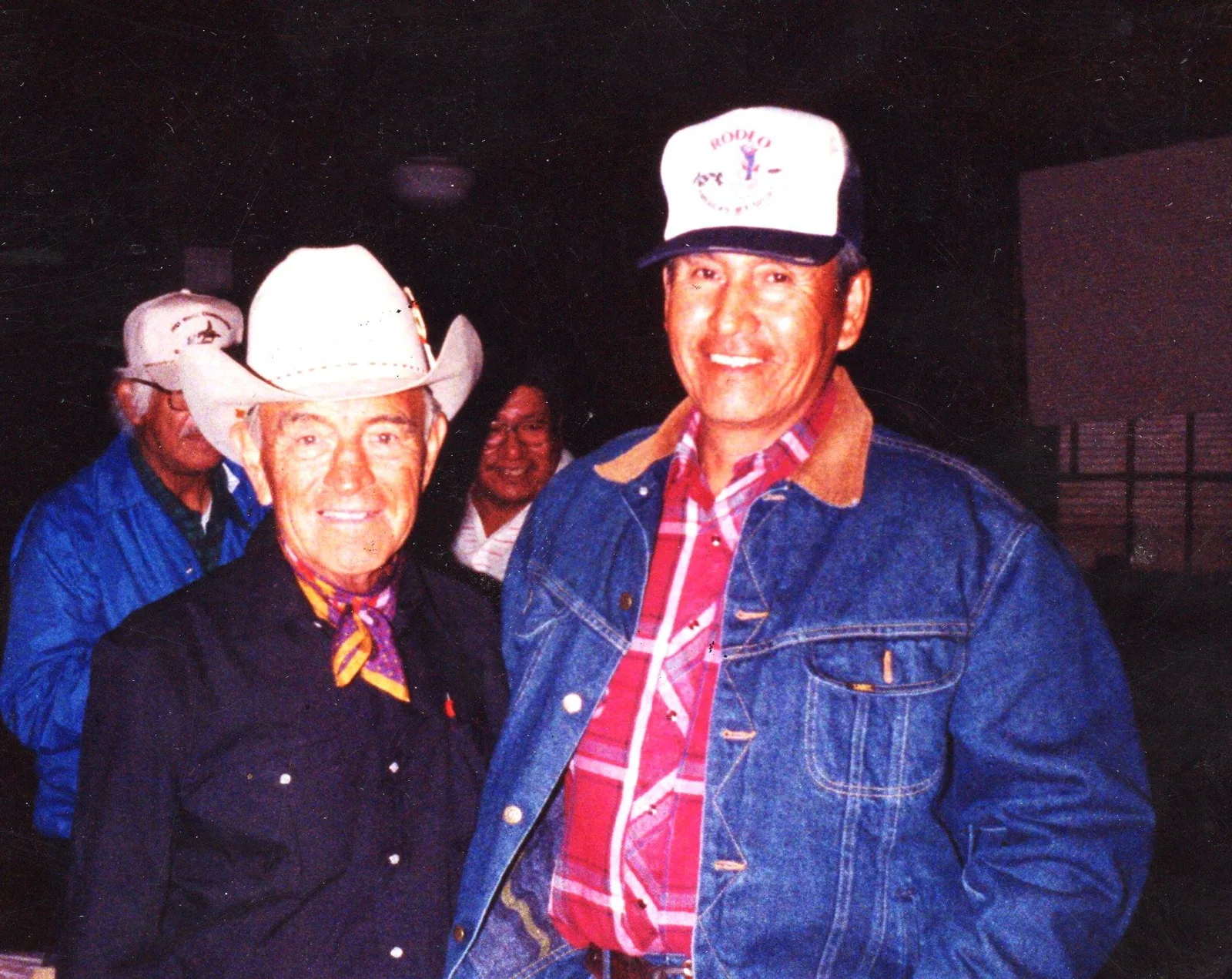 An elderly man in a white cowboy hat and neckerchief stands smiling beside a man in a denim jacket and rodeo cap.