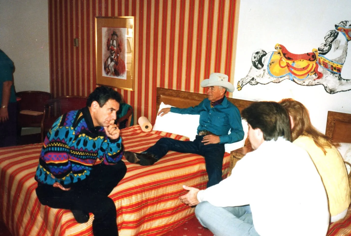 An elder in a cowboy hat reclines on a striped bed in a hotel room, talking with several people seated nearby.
