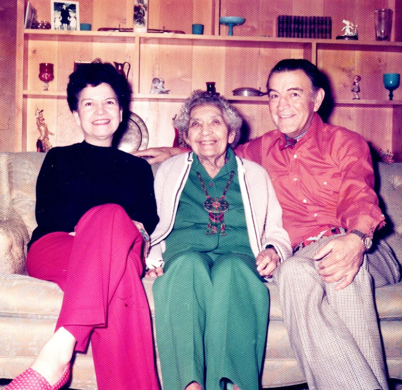A woman, an elderly woman in green with a beaded necklace, and a man in a red shirt sit smiling together on a couch.