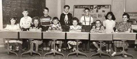 Black-and-white class photo of second graders seated at wooden desks in a classroom with a chalkboard, a teacher beside them; young Phil Lane Jr. stands in back wearing suspenders.