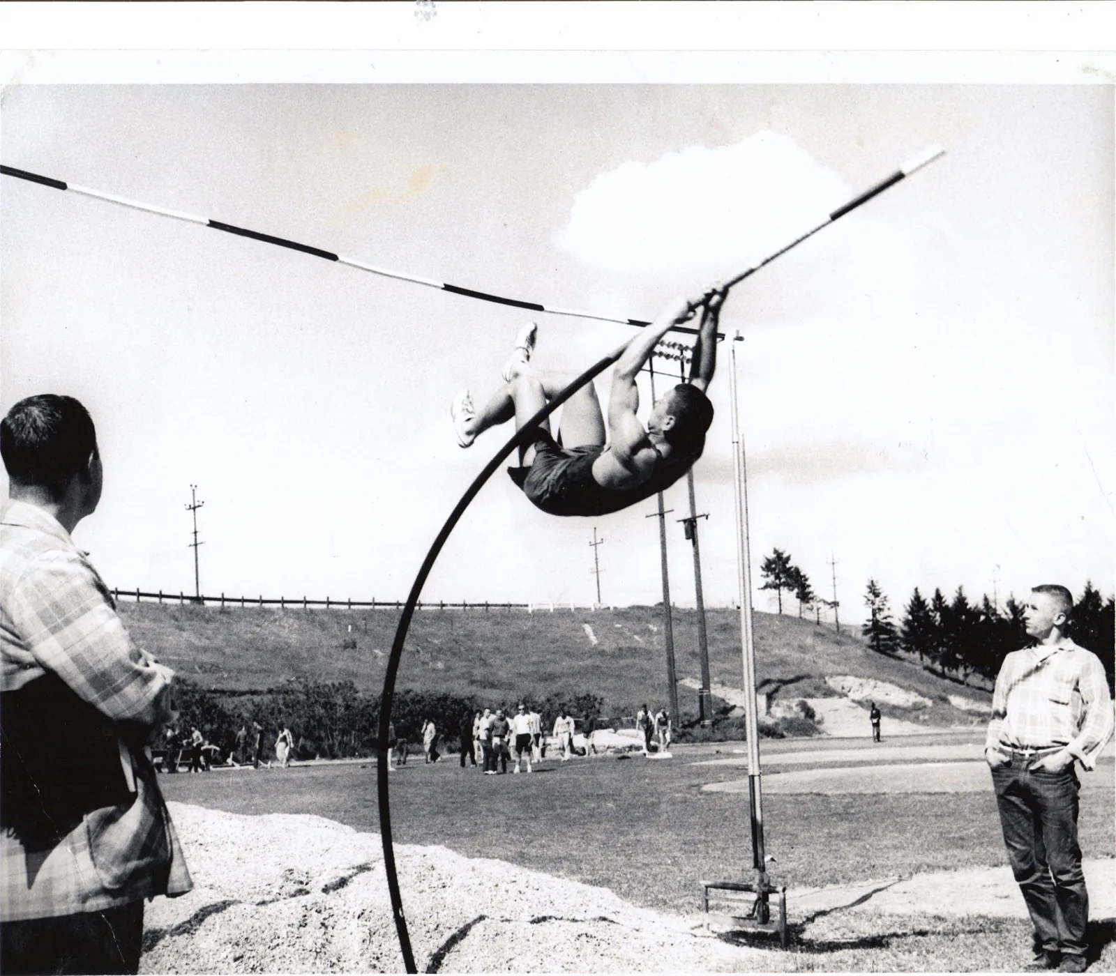 Black-and-white photo of a young athlete mid-vault, curled upside down as a fiberglass pole bends deeply beneath a striped crossbar, while two men watch from the sides of an outdoor track.