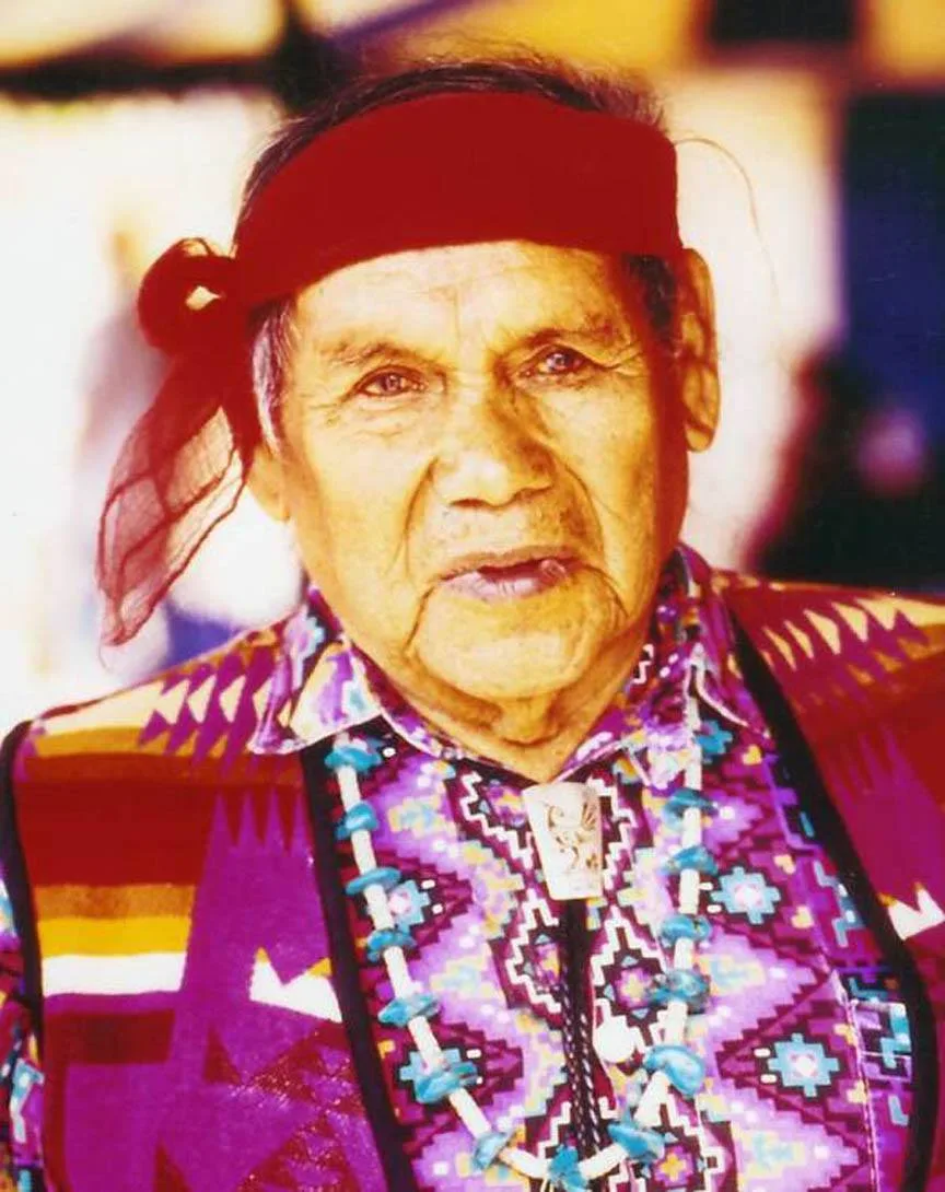 Portrait of an elderly Indigenous man wearing a red headband tied at the side, a turquoise and white bead necklace, a bolo tie, and a vivid purple and pink geometric-patterned shirt and vest.