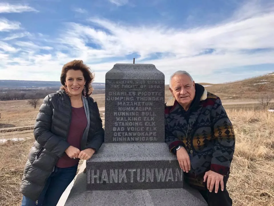 Phil Lane Jr. and his daughter Lisa stand beside a granite monument inscribed 'Ihanktunwan' and listing treaty delegates of 1858, on a grassy hill under a blue sky.