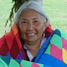 A smiling elder woman with long gray hair, wrapped in a colorful star quilt, seated outdoors on the grass.