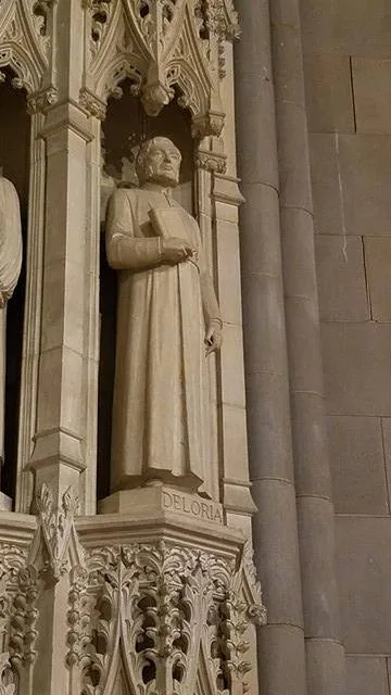 A carved stone statue of a robed man holding a book, standing in a Gothic niche at the National Cathedral, with the name "Deloria" inscribed on its base.