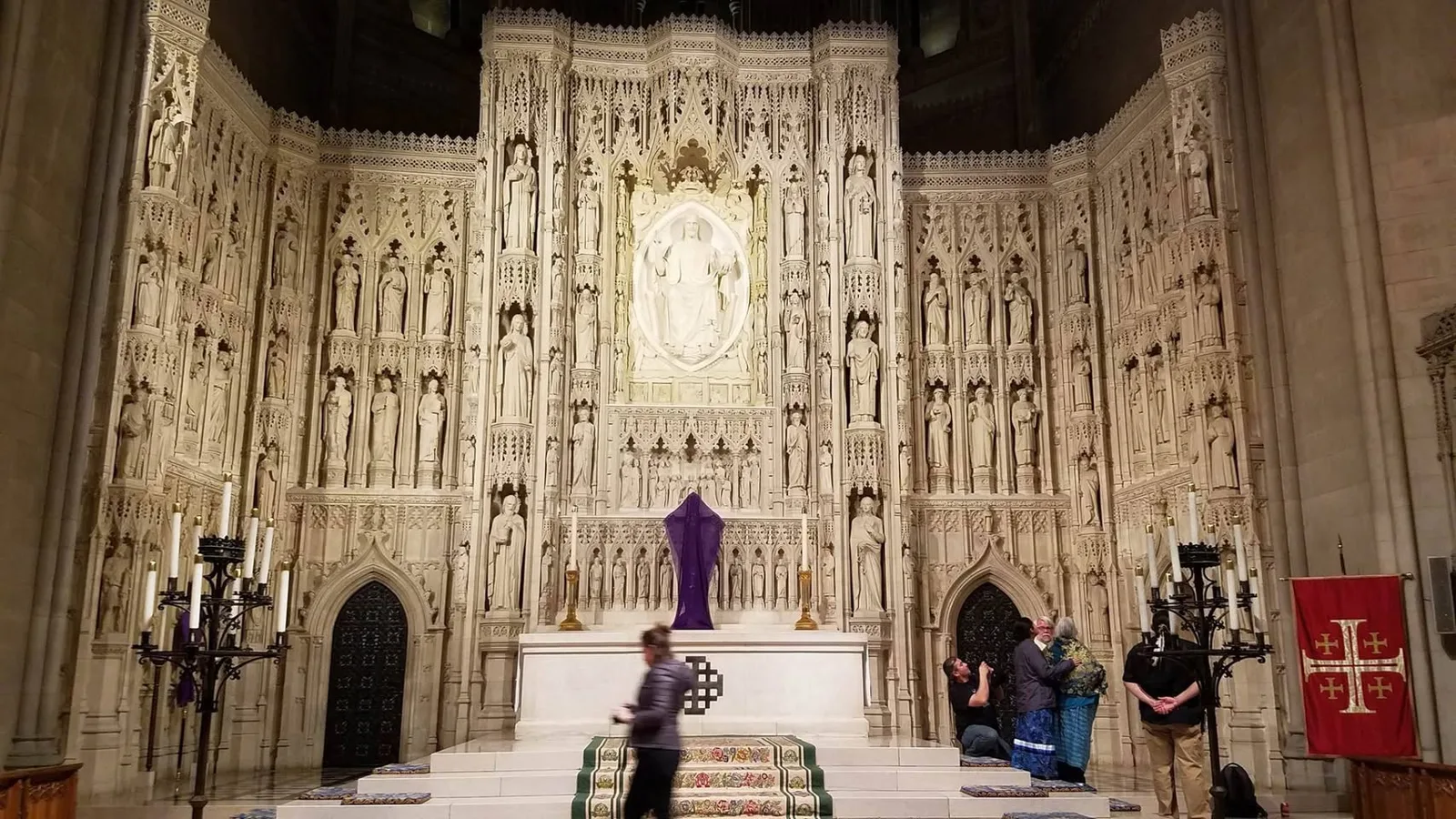 The ornate carved stone High Altar of the National Cathedral, filled with statues of saints, with a small group of people gathered near the altar steps.