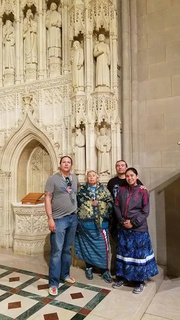 Four relatives, including an elder in a ribbon skirt and a young woman in a ribbon skirt, stand together before carved stone saints at the National Cathedral.