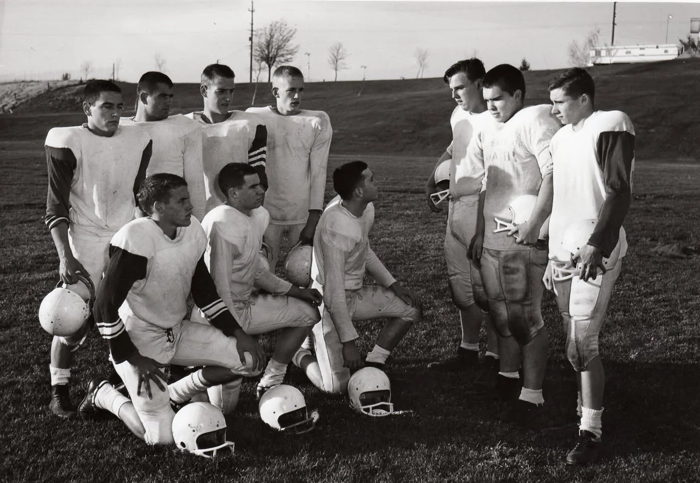 Black-and-white photo of ten young football players in white practice uniforms on a grassy field, some kneeling and some standing, holding their helmets.