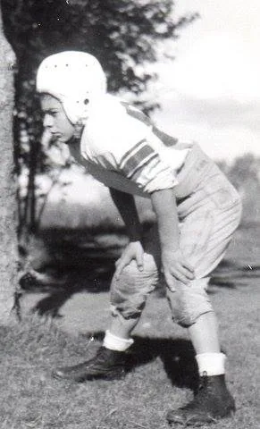 Black-and-white photo of a young football player in a leather helmet and striped jersey, crouched with hands on knees beside a tree.
