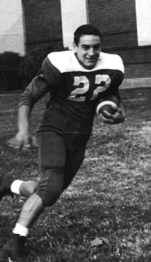 Black-and-white photo of a smiling young football player wearing jersey number 22, running across the grass with a football tucked under his arm.