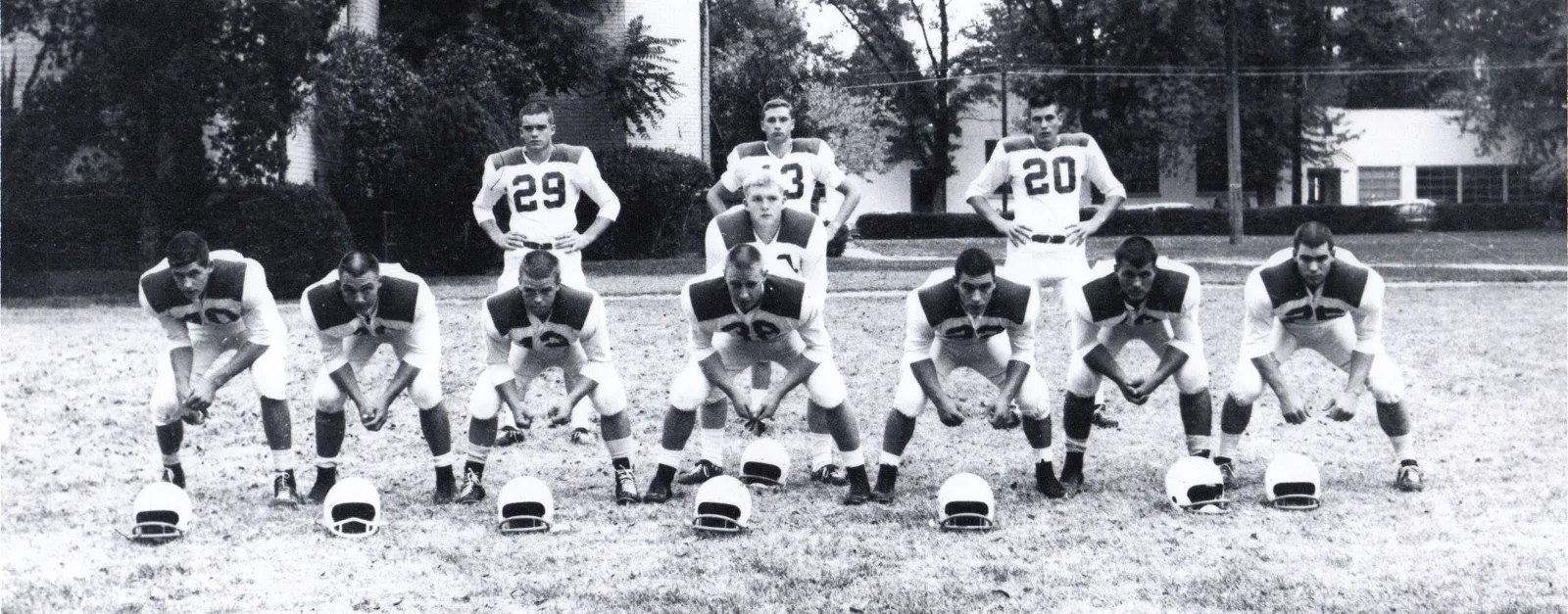 Black-and-white photo of a high school football team posed outdoors, seven linemen crouched in stance with helmets on the ground before them and four backs standing behind.