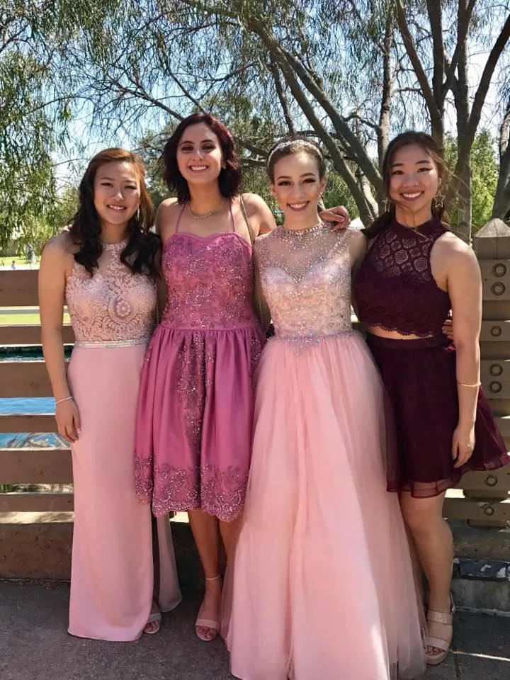 Four smiling young women in formal gowns of pink, rose and burgundy stand arm in arm in front of a wooden fence under sunlit trees.