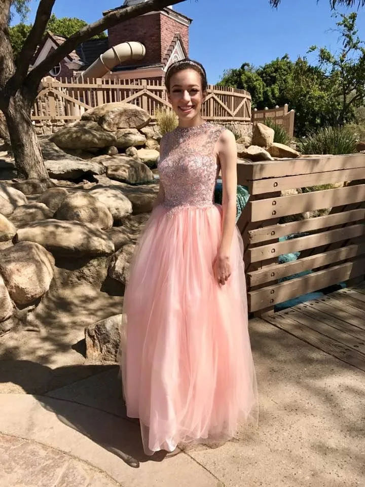 A smiling young woman in a long pink tulle gown with a beaded bodice and a tiara stands on a sunny path beside rocks and a wooden railing.