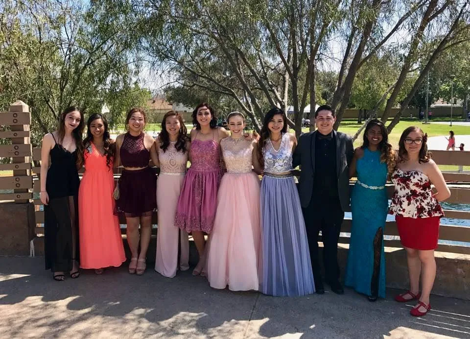 Ten young people in prom attire, nine young women in colorful gowns and one young man in a dark suit, stand in a row in a sunny park.