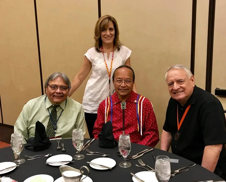 Dr. Bob Whitman, seated in a red ribbon shirt with a bolo tie, at a set banquet table between two smiling men, with Lisa Brown Jordan standing behind him in a white blouse.